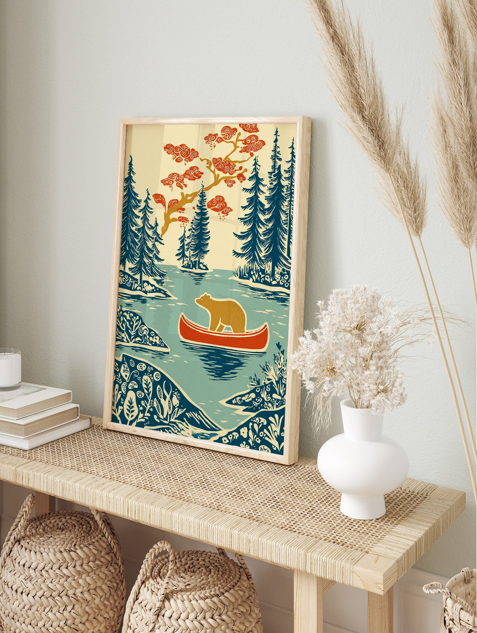 Woodcut style print of a brown bear in a red canoe on a lake, surrounded by blue evergreen, in an alternate framed mockup