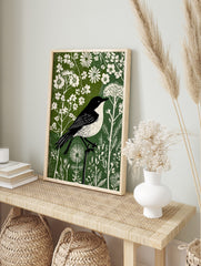 Black and white bird in linocut style perched on branch amidst white flowers and green, in an alternate framed mockup