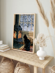 Illustrative print of a black cat sitting before a glowing wood-burning stove with a snowy, in an alternate framed mockup