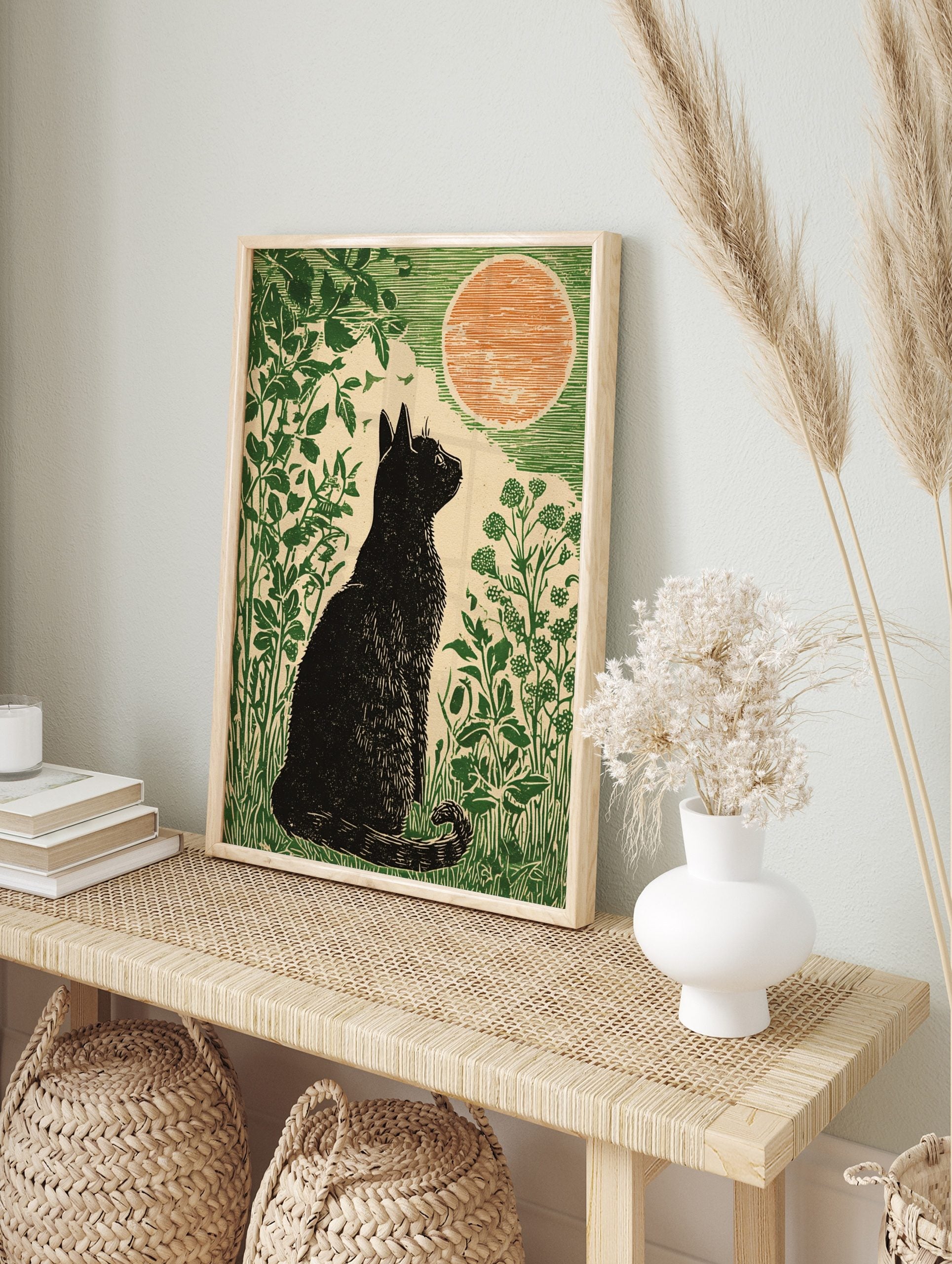 Linocut style art print of a black cat sitting in green foliage, looking at a large orange, in an alternate framed mockup