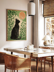 Linocut style art print of a black cat sitting in green foliage, looking at a large orange moon, in a styled room mockup