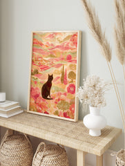 Illustrative print of black cat in colourful landscape with pink hills, green trees, and, in an alternate framed mockup