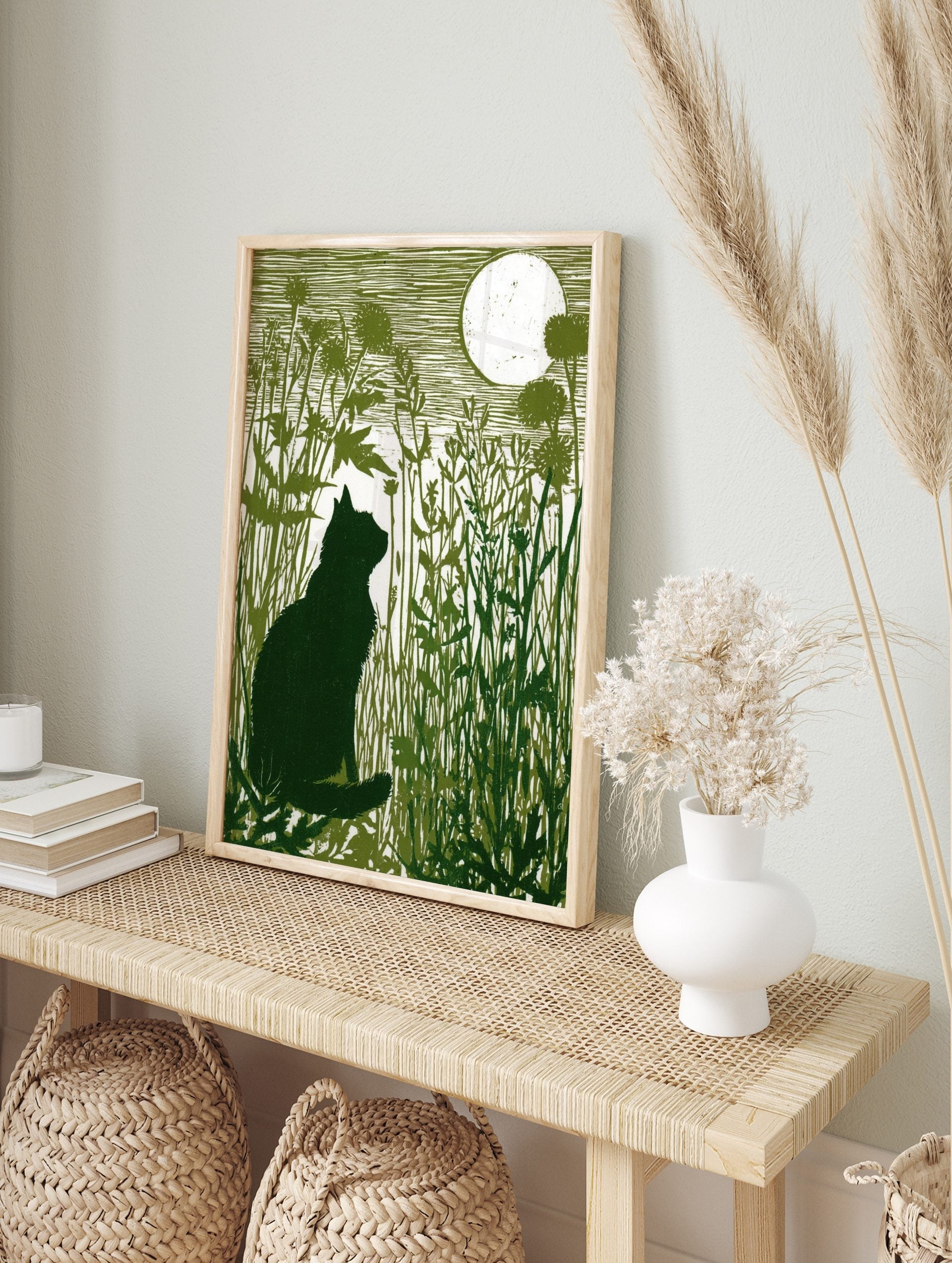 Linocut-style art print of a black cat silhouette sitting in tall green grass, looking at a, in an alternate framed mockup
