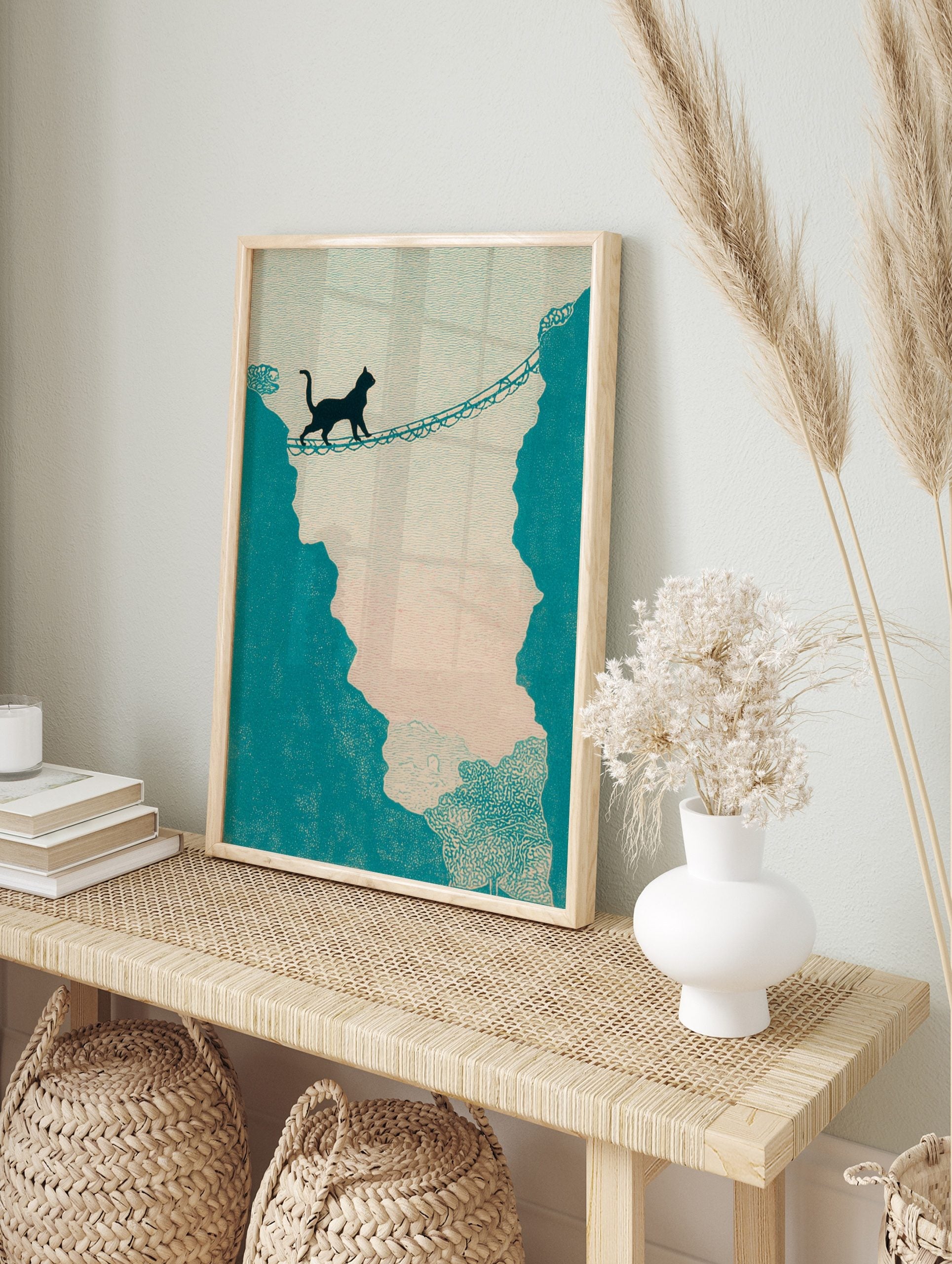 Illustrative print of a black cat walking across a rope bridge between teal cliffs against a, in an alternate framed mockup