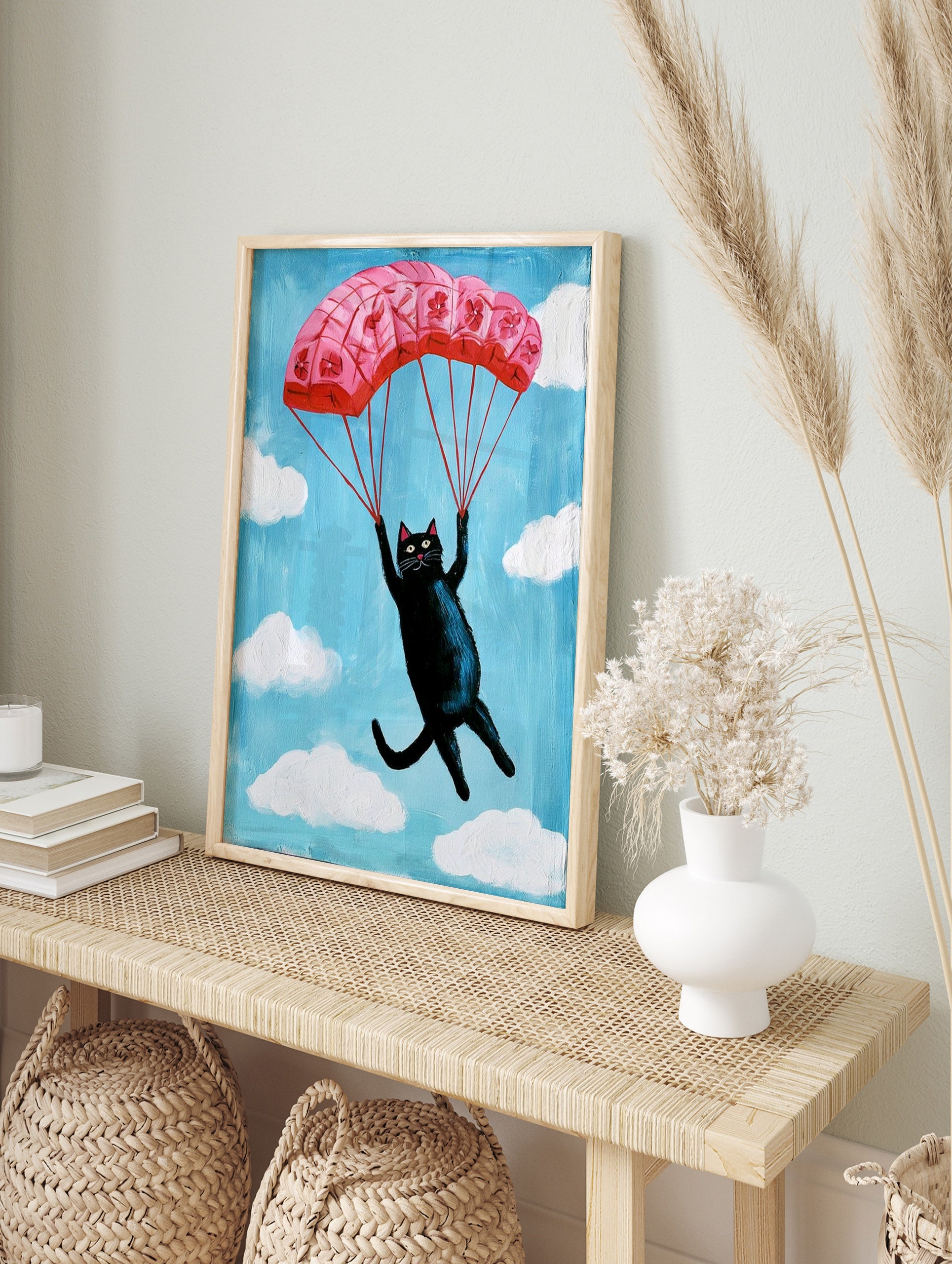 Whimsical illustration of a black cat descending with a pink floral parachute in a blue sky, in an alternate framed mockup