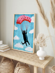 Whimsical illustration of a black cat descending with a pink floral parachute in a blue sky, in an alternate framed mockup