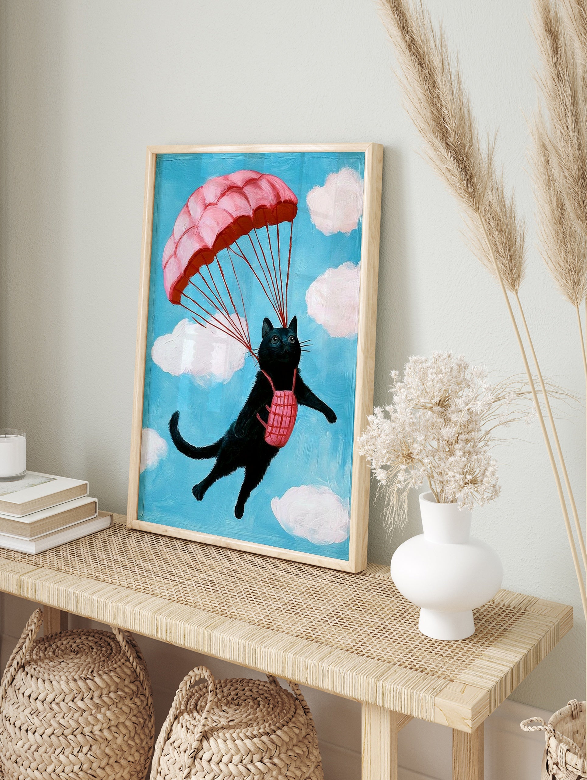 A whimsical illustration of a black cat parachuting with a pink parachute against a blue sky, in an alternate framed mockup