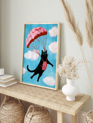 A whimsical illustration of a black cat parachuting with a pink parachute against a blue sky, in an alternate framed mockup