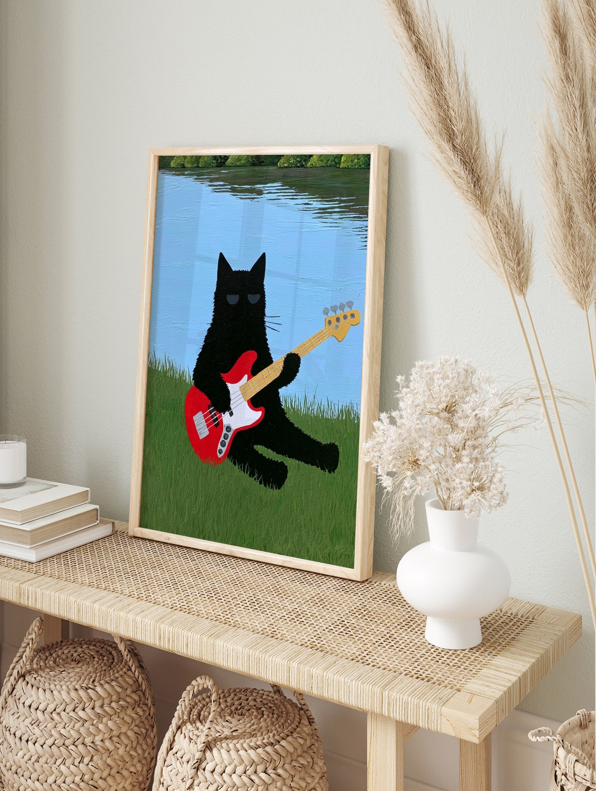 Black cat with sunglasses playing a red bass guitar in green grass by a blue lake, in an alternate framed mockup