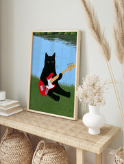 Black cat with sunglasses playing a red bass guitar in green grass by a blue lake, in an alternate framed mockup
