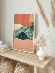 Illustrative print of a black cat with green eyes peeking from under a green palm leaf, in an alternate framed mockup