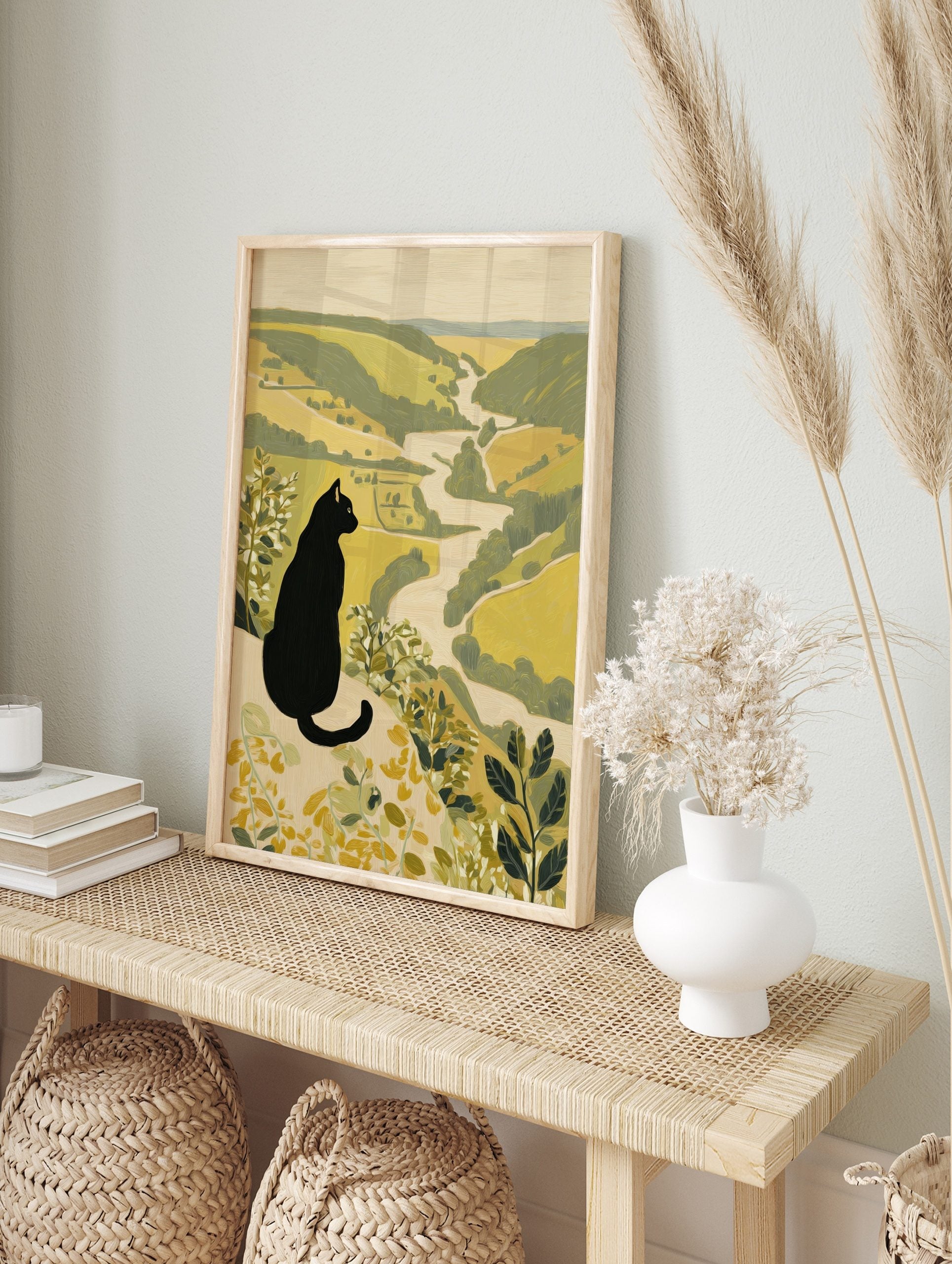 Illustrative print of a black cat with yellow eyes overlooking a green and beige river valley, in an alternate framed mockup
