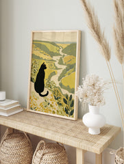 Illustrative print of a black cat with yellow eyes overlooking a green and beige river valley, in an alternate framed mockup