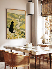 Illustrative print of a black cat with yellow eyes overlooking a green and beige river valley, in a styled room mockup