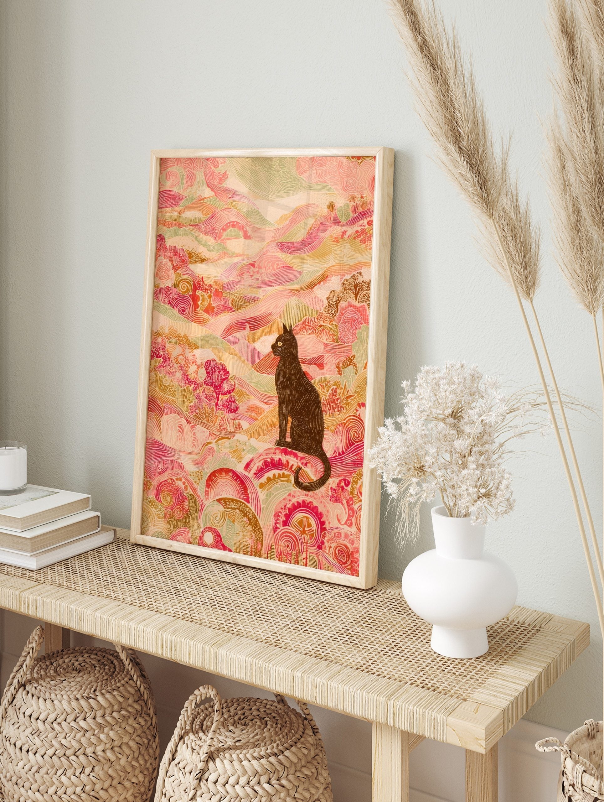 Illustrative black cat with yellow eyes in an ornate pink and green patterned landscape art, in an alternate framed mockup