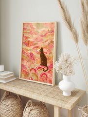 Illustrative black cat with yellow eyes in an ornate pink and green patterned landscape art, in an alternate framed mockup
