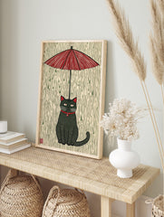 Stylised black cat with red collar and eyes sitting under a red umbrella in green rain, in an alternate framed mockup