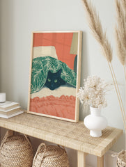 Illustrative print of a black cat with green eyes peeking from under a green tropical leaf, in an alternate framed mockup