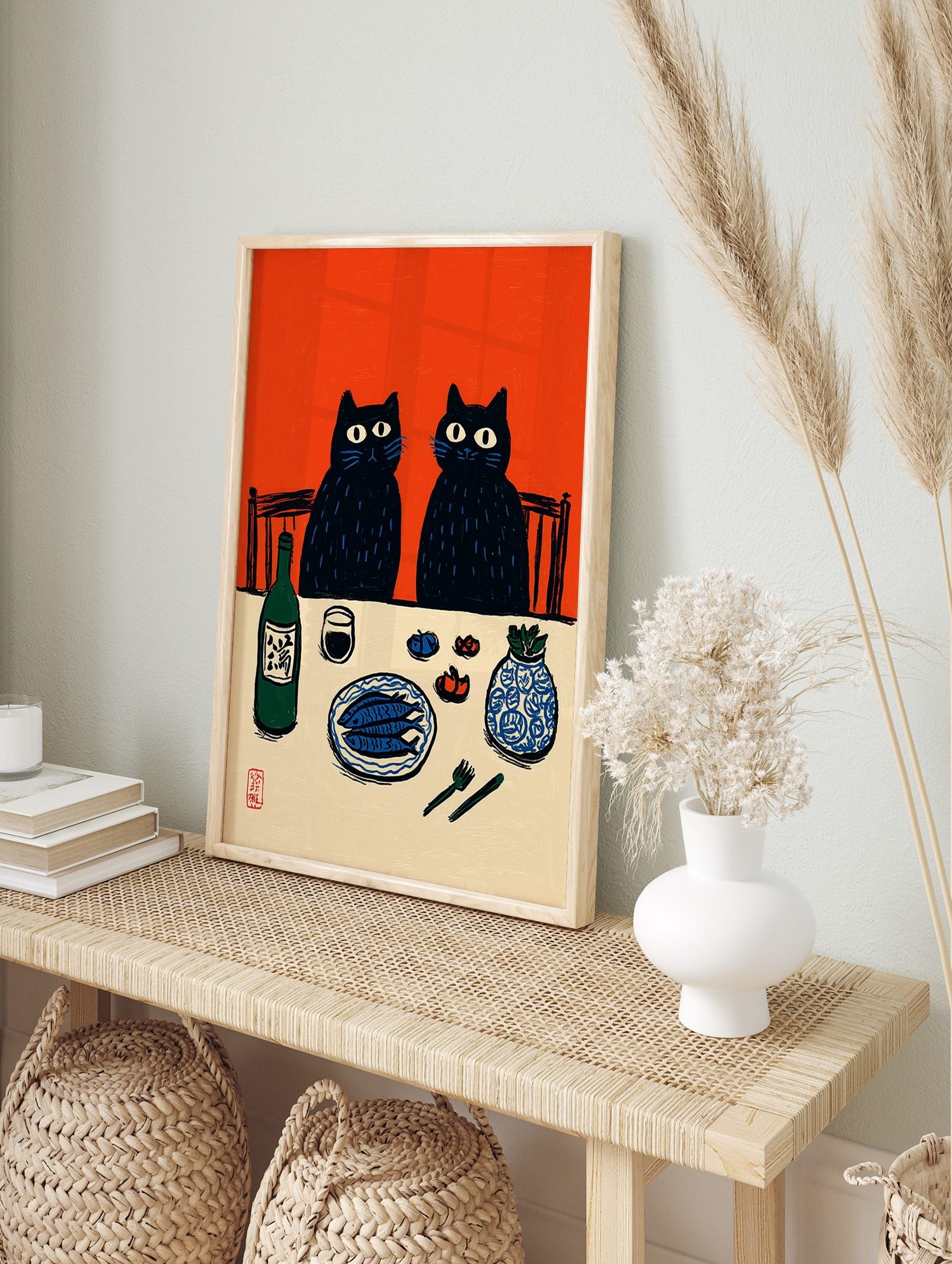 Illustrative print of two black cats at a dinner table with fish, sake, and a vase, against, in an alternate framed mockup