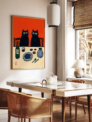 Illustrative print of two black cats at a dinner table with fish, sake, and a vase, against an, in a styled room mockup