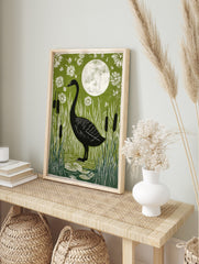 Black goose standing in a pond with tall white reeds, black cattails, a full moon, and white, in an alternate framed mockup