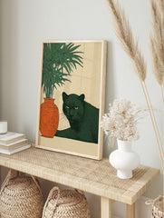 Illustrative black panther lying next to an ornate orange vase and green plant on a beige, in an alternate framed mockup