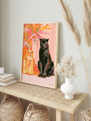 Black panther sitting beside a vase of vibrant orange and pink tropical leaves on a pink, in an alternate framed mockup