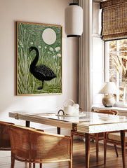 Black swan standing in water surrounded by green cattails and white flowers, with a large white moon, in a styled room mockup
