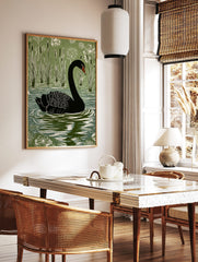 Black swan linocut style art print swimming in a lake with green reeds and white flowers, in a styled room mockup