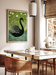 Black swan linocut print on water with green botanical foliage and white flowers in background, in a styled room mockup