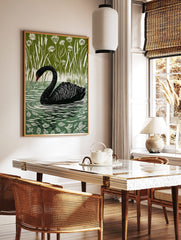 Black swan swimming in water with white ripples, surrounded by tall green reeds and white flowers, in a styled room mockup