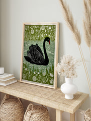 Black swan swimming on water, surrounded by green reeds and white flowers, in a linocut-style, in an alternate framed mockup