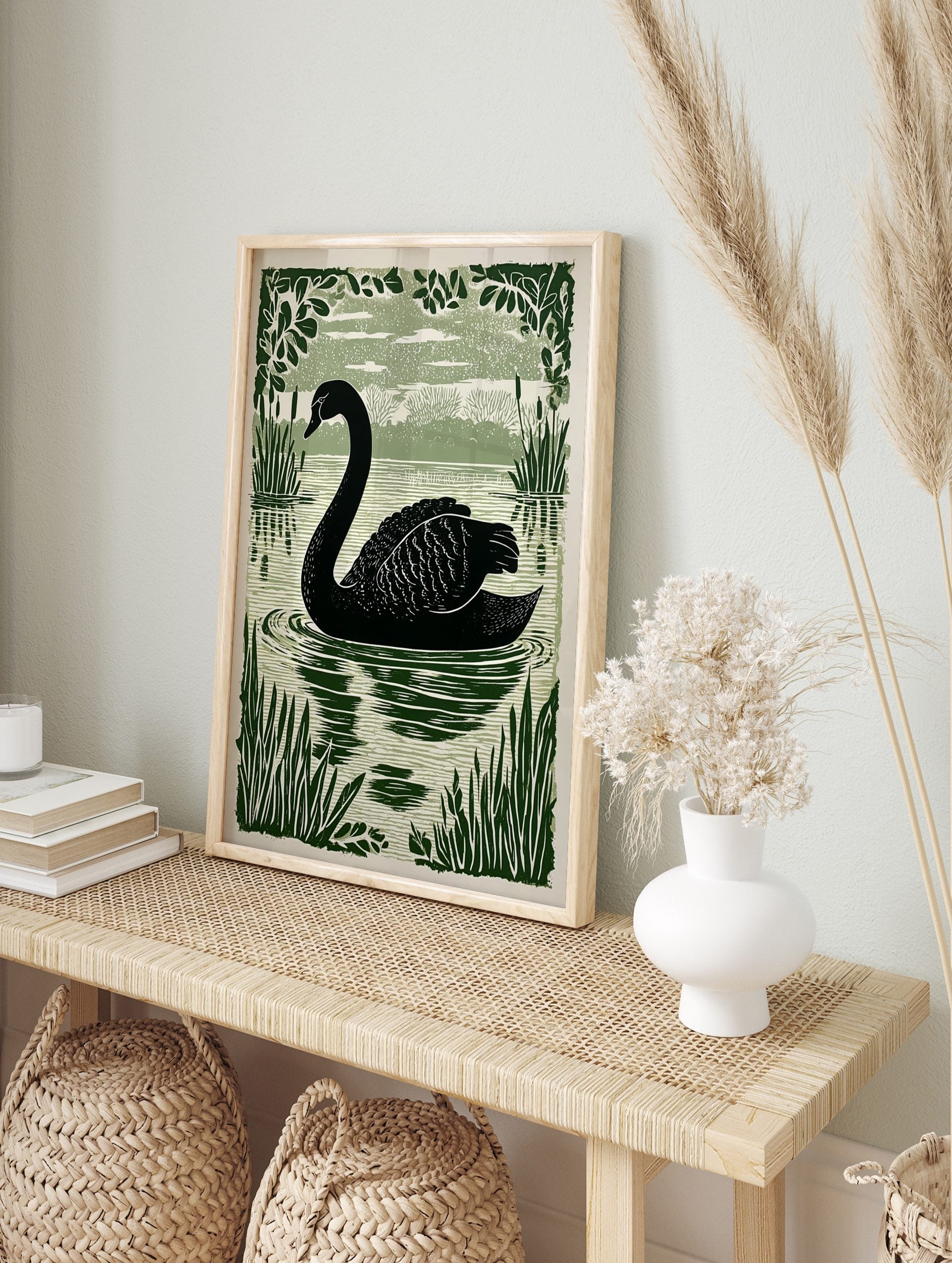 Black swan linocut print swimming in a lake with dark green reeds and foliage, light green, in an alternate framed mockup