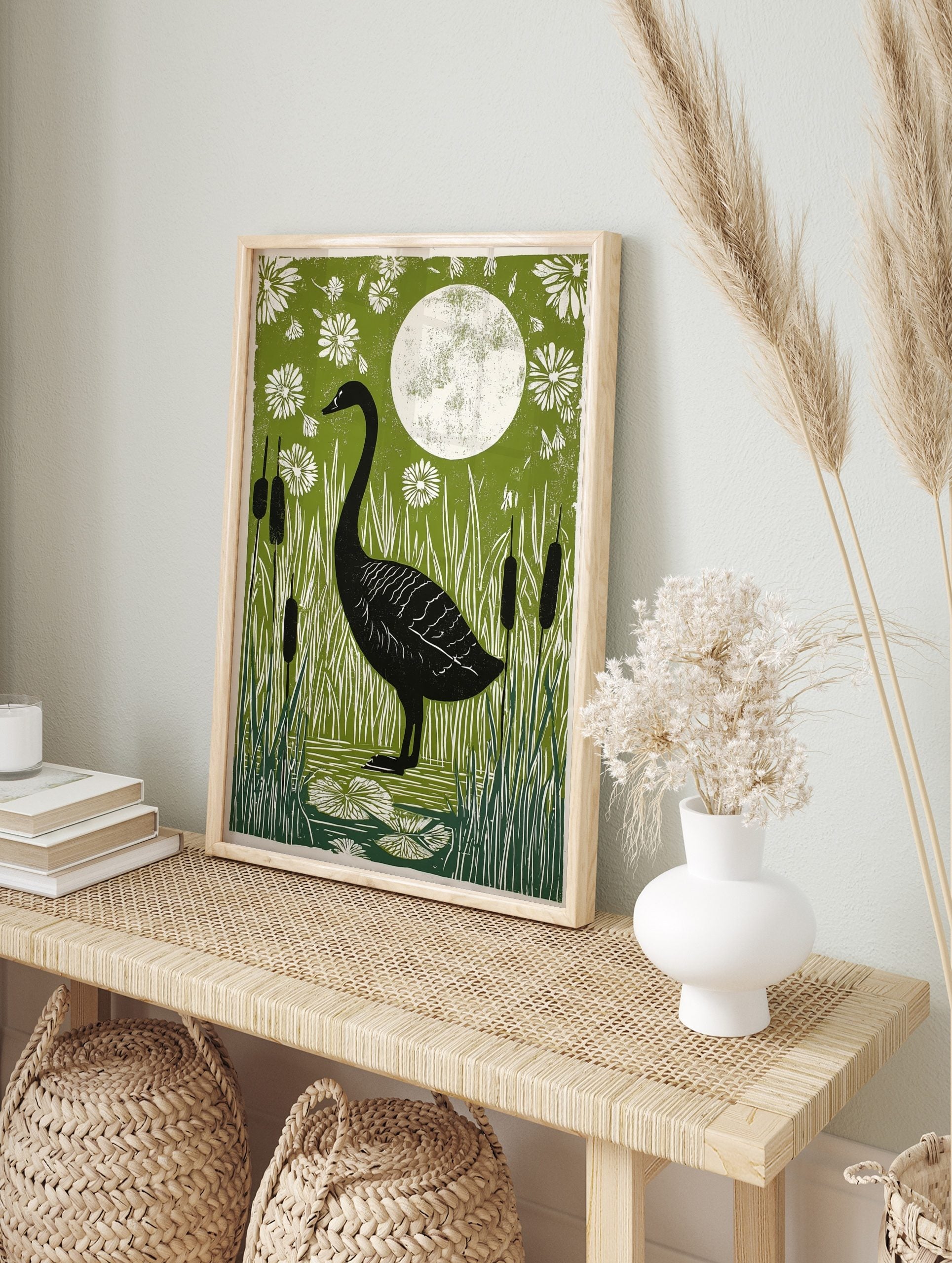 Black swan standing in a pond with white reeds, lily pads, and flowers under a full moon on a, in an alternate framed mockup
