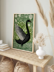Black swan with outstretched wing swimming in water, surrounded by white wildflowers and, in an alternate framed mockup