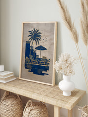 Blue and gold illustrative art print of a palm tree, deck chairs, and parasol by a pool on, in an alternate framed mockup