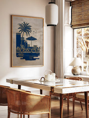 Blue and gold illustrative art print of a palm tree, deck chairs, and parasol by a pool on beige, in a styled room mockup