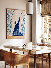 Illustrative print of a woman in a blue outfit in a kneeling yoga pose, reaching for blue flowers, in a styled room mockup