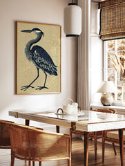 Illustrative blue heron standing on a textured beige background with subtle gold botanical elements, in a styled room mockup