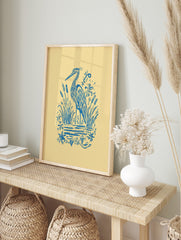 Blue illustrative heron standing among cattails and water plants on a light yellow background, in an alternate framed mockup