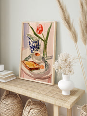 Watercolor painting of a breakfast table with a red tulip, blue-and-white jug, toast, and egg, in an alternate framed mockup