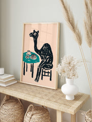 Whimsical black camel sitting at a teal table with cake and a cup, against a pale pink, in an alternate framed mockup