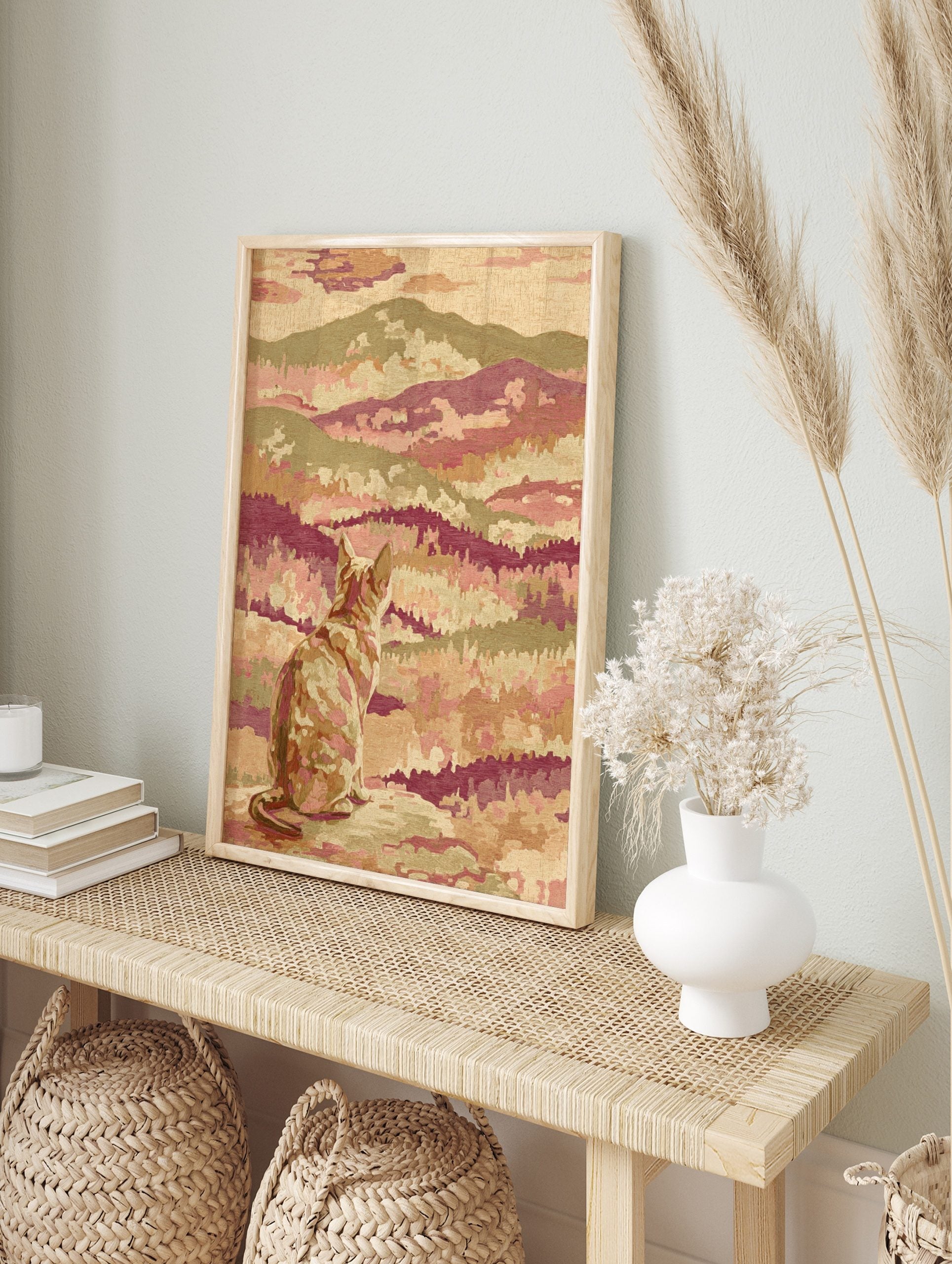 Stylised cat looking at a layered mountain landscape print with tapestry texture in pink, in an alternate framed mockup