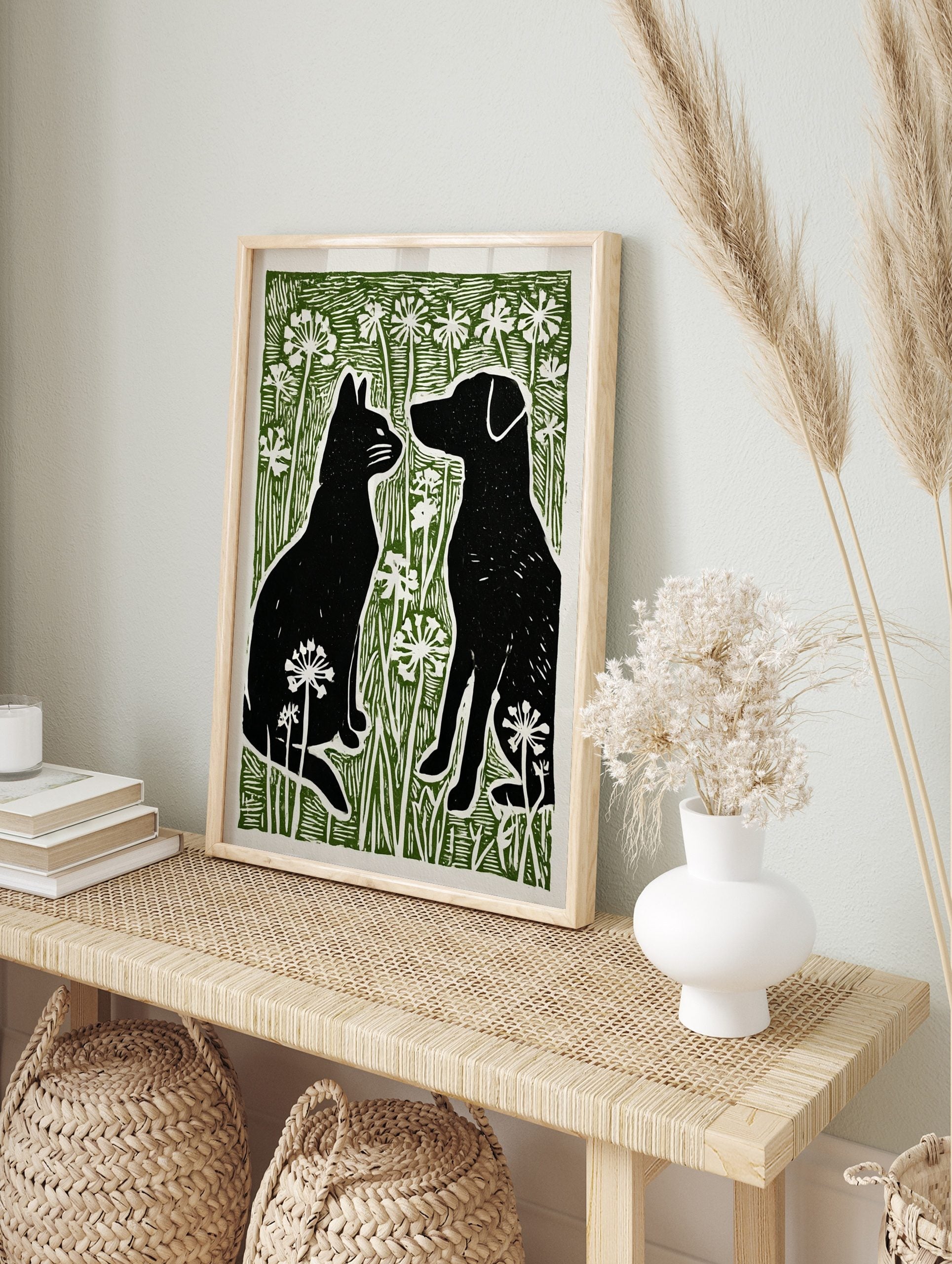 Black cat and dog silhouettes facing each other in green grass with white flowers, linocut, in an alternate framed mockup