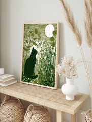 Green linocut print of a black cat silhouette in tall grass looking at a full moon, in an alternate framed mockup