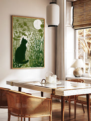 Green linocut print of a black cat silhouette in tall grass looking at a full moon, in a styled room mockup