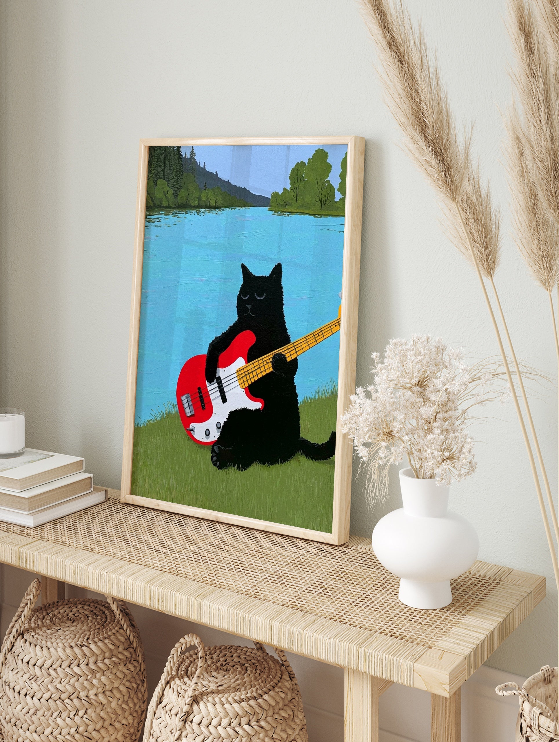 Black cat playing a red electric bass guitar on a grassy riverbank with a blue river and, in an alternate framed mockup
