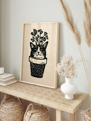 Linocut style illustration of a cat face emerging from a plant pot with clover leaves, black, in an alternate framed mockup