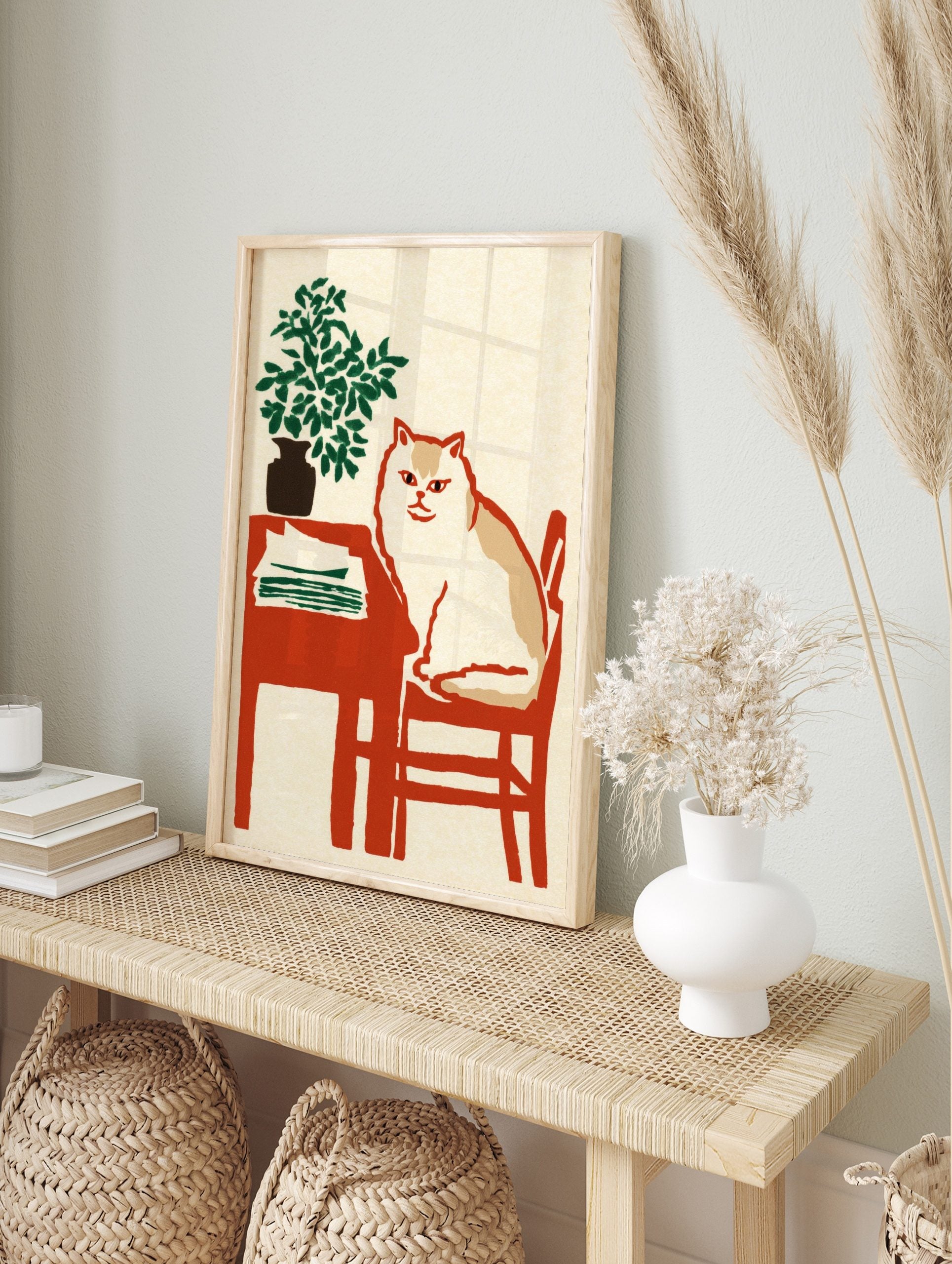 Illustrative print of a white and tan cat on a red chair next to a table with a green plant, in an alternate framed mockup