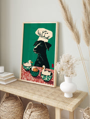 Illustrative wall art print of a black dog in a chef's hat with two ducks and eggs on a red, in an alternate framed mockup
